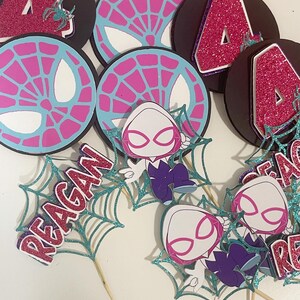 May include: A collection of Spider-Gwen themed party decorations. Includes black circular cutouts with a pink glitter number 4, and spiderweb designs. Also features cutouts of Spider-Gwen and the name "Reagan" in pink glitter.