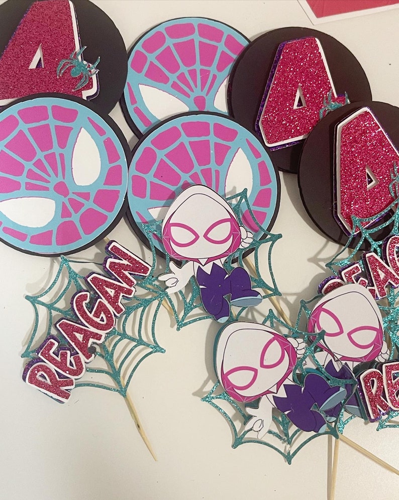 May include: A collection of Spider-Gwen themed party decorations. Includes black circular cutouts with a pink glitter number 4, and spiderweb designs. Also features cutouts of Spider-Gwen and the name "Reagan" in pink glitter.