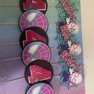 May include: A set of handmade cupcake toppers featuring a Spider-Gwen theme. The toppers include the number 4 in glittery pink, spiderweb designs, and the name "Reagan" in a matching font. The toppers are attached to wooden skewers.