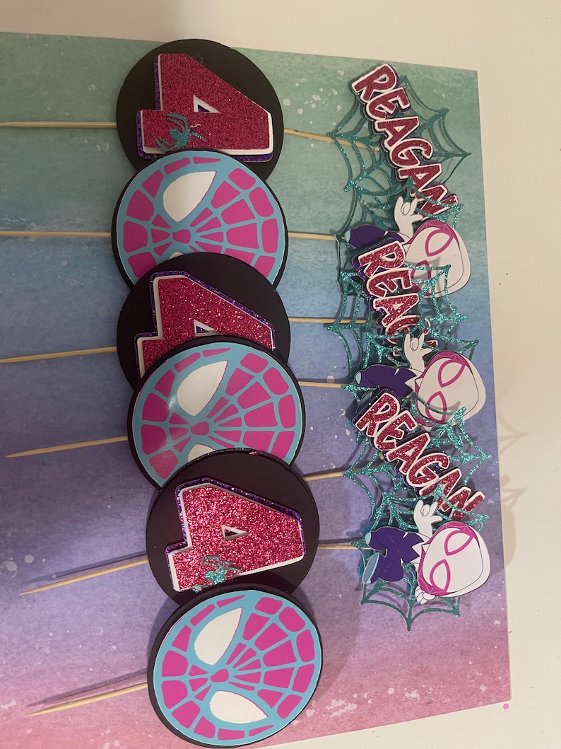 May include: A set of handmade cupcake toppers featuring a Spider-Gwen theme. The toppers include the number 4 in glittery pink, spiderweb designs, and the name "Reagan" in a matching font. The toppers are attached to wooden skewers.