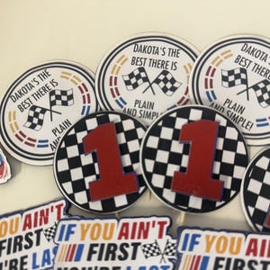 May include: A collection of racing-themed party decorations. Several circular decorations feature checkered flags and the text "DAKOTA'S THE BEST THERE IS" and "PLAIN AND SIMPLE!". Other decorations include a large red number 1 on a checkered background and text that reads "IF YOU AIN'T FIRST YOU'RE LAST".