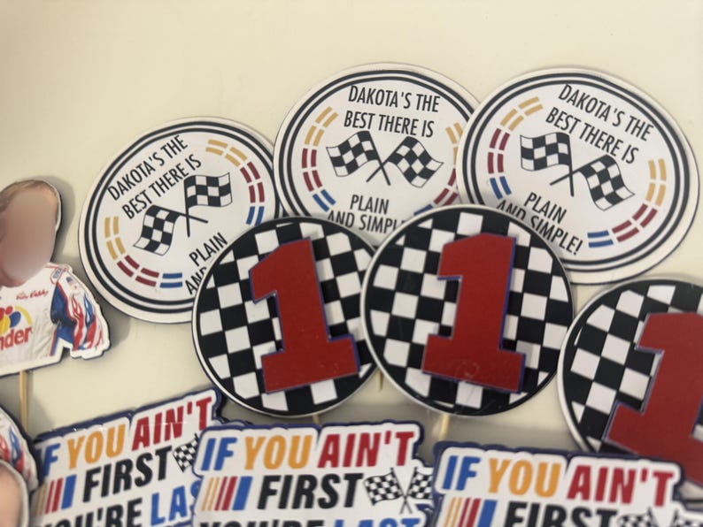 May include: A collection of racing-themed party decorations. Several circular decorations feature checkered flags and the text "DAKOTA'S THE BEST THERE IS" and "PLAIN AND SIMPLE!". Other decorations include a large red number 1 on a checkered background and text that reads "IF YOU AIN'T FIRST YOU'RE LAST".
