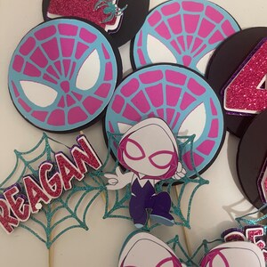 May include: A collection of party decorations featuring a Spider-Gwen theme. Includes circular toppers with a spiderweb design in pink and blue, and the number 4 in glitter. Also includes a teal glitter web with the name "REAGAN".