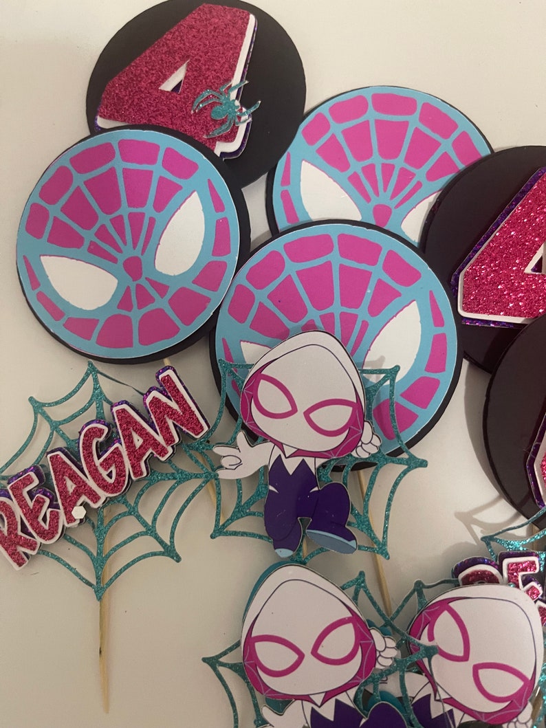 May include: A collection of party decorations featuring a Spider-Gwen theme. Includes circular toppers with a spiderweb design in pink and blue, and the number 4 in glitter. Also includes a teal glitter web with the name "REAGAN".