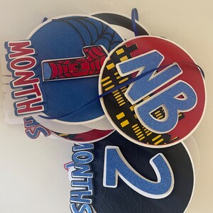 Puede incluir: Un conjunto de decoraciones redondas y superpuestas con tem&aacute;tica de superh&eacute;roes. Las decoraciones presentan los n&uacute;meros 1 y 2, y las palabras "MONTHS" y "NB" en azul y rojo. Tambi&eacute;n se ven telara&ntilde;as y paisajes urbanos.