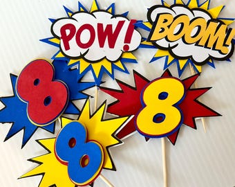 May include: Superhero-themed party decorations featuring speech bubbles with "POW!" and "BOOM!" text, along with number "8" cutouts. The decorations are in red, yellow, blue, and white, attached to wooden sticks for easy placement.