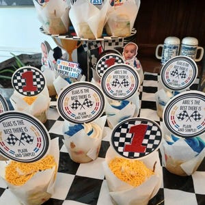 May include: Cupcakes with racing-themed toppers. The toppers feature a checkered flag design, the number 1, and the phrases "Abel's the best there is" and "Elias is the best there is, plain and simple!" The cupcakes are on a black and white checkered surface.