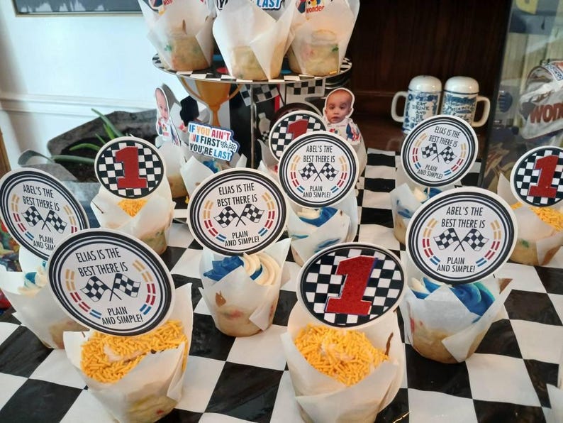May include: Cupcakes with racing-themed toppers. The toppers feature a checkered flag design, the number 1, and the phrases "Abel's the best there is" and "Elias is the best there is, plain and simple!" The cupcakes are on a black and white checkered surface.