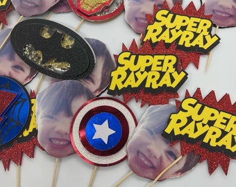May include: A collection of superhero-themed party decorations. The decorations include images of a smiling child, superhero logos, and the text "SUPER RAYRAY" in yellow and black on a red glitter background. The logos include Batman, Captain America, and Iron Man.