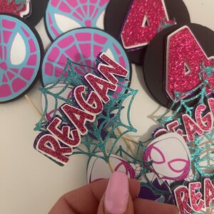 May include: A collection of handmade party decorations featuring a Spider-Gwen theme. The decorations include circular toppers with spiderweb designs, the number "4" in pink glitter, and the name "Reagan" in glitter. The color scheme is pink, blue, and black.