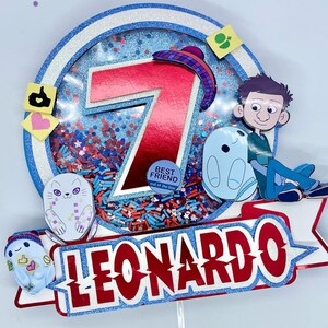 May include: A colorful birthday cake topper with the name "LEONARDO" in red and white. The topper features a large red number 7, cartoon characters, and confetti inside a blue and silver glitter circle.