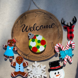 May include: A wooden Christmas door hanger with the word "Welcome" painted on it. The hanger is decorated with a variety of Christmas-themed ornaments, including a gingerbread person, a snowman, a candy cane, a snowflake, and a reindeer. The ornaments are painted in various colors, including red, green, blue, white, and brown.