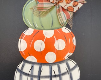 May include: A stack of three painted pumpkins with a plaid bow. The top pumpkin is green, the middle pumpkin is orange with white polka dots, and the bottom pumpkin is white with black plaid.