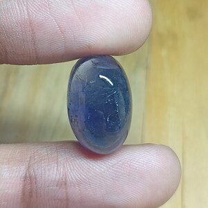 May include: A smooth, oval-shaped gemstone with a deep blue color. The stone has a slight purple hue and is translucent.