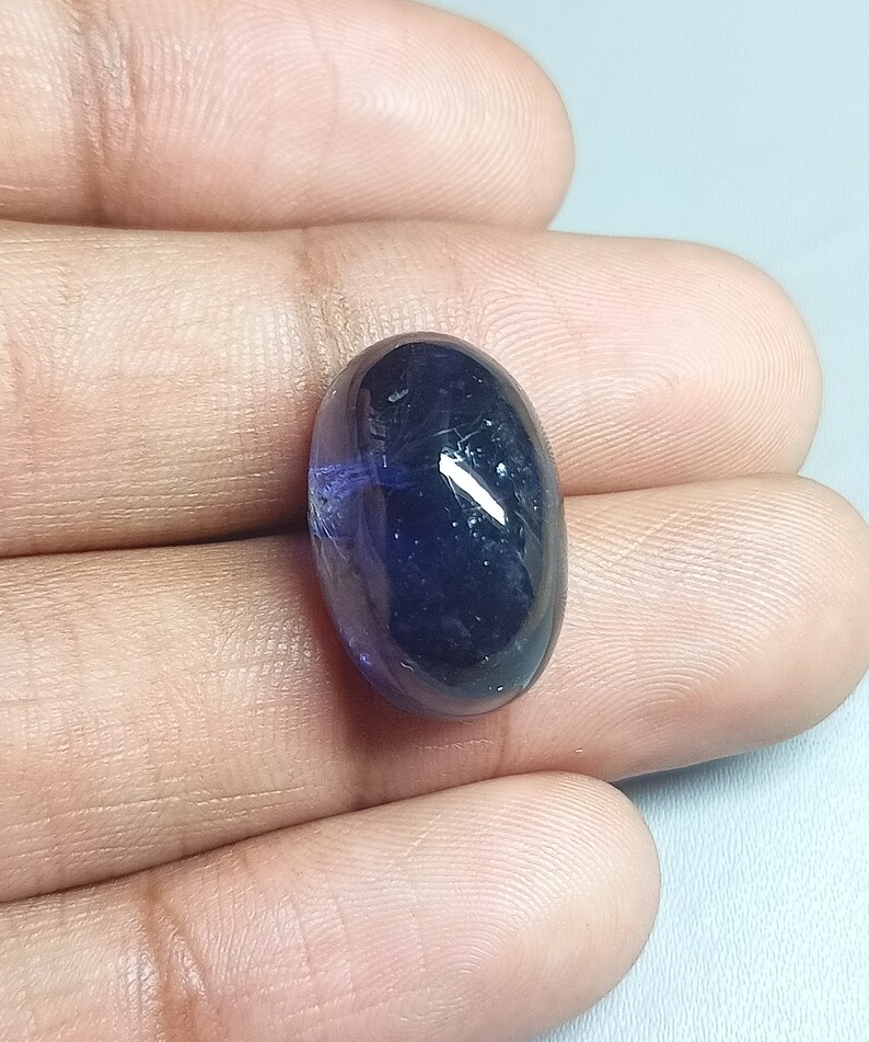 May include: A dark blue oval-shaped gemstone with a slight purple hue. The gemstone is smooth and polished.