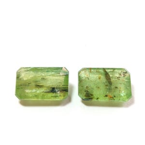 May include: Two green rectangular gemstones with inclusions. The gemstones are cut into a rectangular shape with beveled edges.