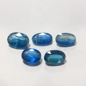 May include: Five oval-shaped blue gemstones with a light blue hue. The gemstones are faceted and have a slight shimmer.