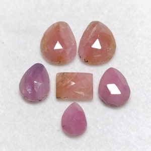 Puede incluir: Seis gemas rosadas de varias formas y tamaños, incluyendo cortes en forma de lágrima, rectangulares y cuadrados. Las gemas están dispuestas en un patrón de flor en una superficie blanca.