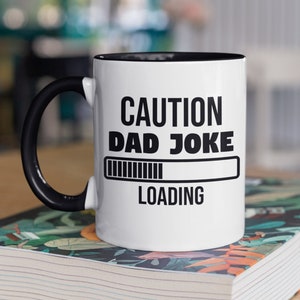 May include: White coffee mug with a black rim and handle. The mug has black text that reads "CAUTION DAD JOKE LOADING" with a progress bar underneath.
