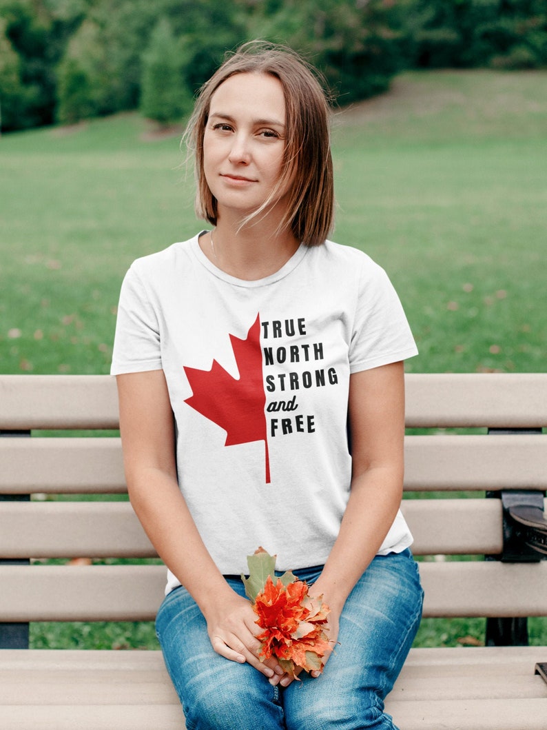 May include: A woman wearing a white t-shirt with a red maple leaf and the text "TRUE NORTH STRONG and FREE". She is sitting on a wooden bench and holding a bouquet of orange flowers.
