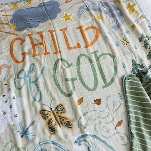 以下が含まれることがあります： オレンジと緑の文字で「A Child of God」と書かれた、薄い緑とベージュのモスリン製ベビーブランケット。ブランケットには、太陽、雲、蝶、蜂など、さまざまなイラストが描かれています。