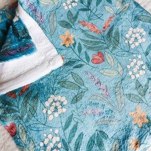 May include: A soft, blue fleece blanket with a white backing. The blanket features a floral pattern with green leaves, red, yellow, and white flowers.
