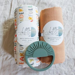 May include: Two muslin swaddle blankets, one with a colorful alphabet print and one in a light brown color, with a green silicone teether shaped like a sun. Both blankets have a circular label with the text "Little Genesis Co. Muslin Swaddle Blanket".