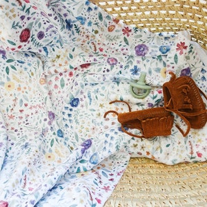 Op de afbeelding: Een witte muslin-wikkeldeken met een bloemenprint in tinten roze, blauw, geel en groen. De deken is gevouwen en geplaatst in een geweven mand met een paar bruine leren baby-moccasins en een groene speen.