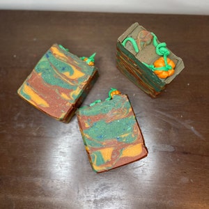 Puede incluir: Tres barras de jabón hechas a mano con un patrón de remolino verde, marrón y naranja. Cada barra está coronada con una pequeña calabaza naranja y un tallo verde.