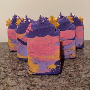 May include: Five bars of handmade soap with a swirl of pink, purple, and gold. Each bar has a decorative purple and gold swirl on top.