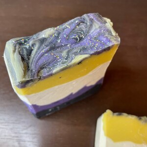 Könnte beinhalten: Eine mehrfarbige Seife mit einem Wirbelmuster. Die Seife hat Schichten in Lila, Schwarz, Weiß und Gelb. Die oberste Schicht hat einen lila und schwarzen Wirbel mit Glitzer.
