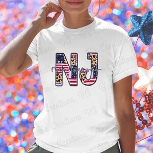 May include: White t-shirt with the letters "NJ" in a patriotic design. The "N" features an American flag pattern, the "J" has a leopard print, and the text "New Jersey" is written in cursive. Decorative stars are in the background.