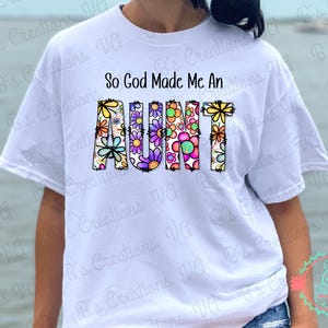 Peut inclure: T-shirt blanc avec le texte "So God Made Me An AUNT" en lettres colorées décorées de fleurs.
