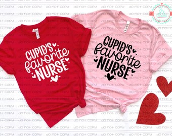 以下が含まれることがあります： 赤とピンクの2枚のTシャツ。それぞれに「Cupid's Favorite Nurse」の文字が白と黒でプリントされています。デザインにはハートが含まれています。赤いハートの切り抜きも2つ見えます。