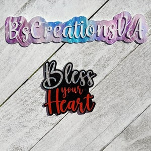 May include: A black and red glitter sign with the text "Bless your heart". The sign is on a white wooden background.