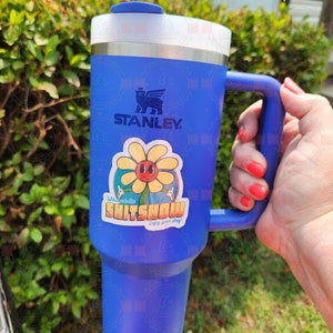 K&ouml;nnte beinhalten: Eine blaue Stanley-Reisebecher mit einem Aufkleber, auf dem "Willkommen im Shitshow Genie&szlig;e deinen Aufenthalt!" steht, und einer Cartoon-Blume mit einem Gesicht.