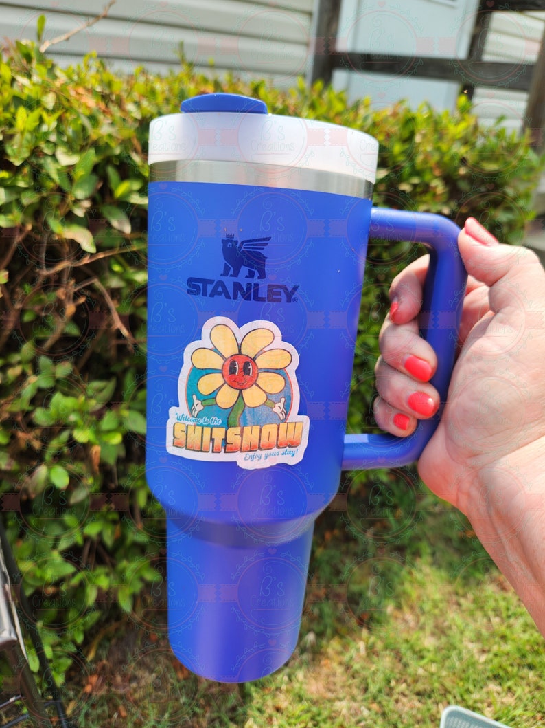 K&ouml;nnte beinhalten: Eine blaue Stanley-Reisebecher mit einem Aufkleber, auf dem "Willkommen im Shitshow Genie&szlig;e deinen Aufenthalt!" steht, und einer Cartoon-Blume mit einem Gesicht.