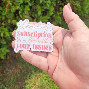 May include: A sticker with the text "Cancel my subscription I'm done with your Issues" in a mix of coral and white colors. The sticker has a white border and is held in a hand against a blurred green background.