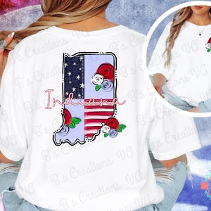 May include: White t-shirt with a graphic of the state of Indiana, incorporating a flag design and floral details. The word "Indiana" is written in a cursive style. The shirt has a round neck and short sleeves.