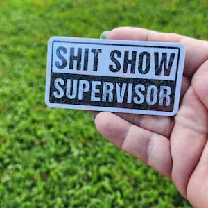 May include: A white sticker with black text that reads "Shit Show Supervisor". The sticker has a glittery texture.