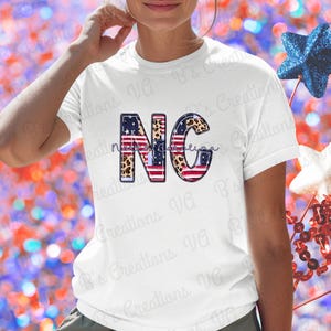 Puede incluir: Camiseta blanca con las letras "NC" en un diseño patriótico, con la bandera estadounidense y estampados de leopardo. La palabra "Carolina" está escrita en cursiva. Decoraciones de estrellas rojas, blancas y azules en el fondo.