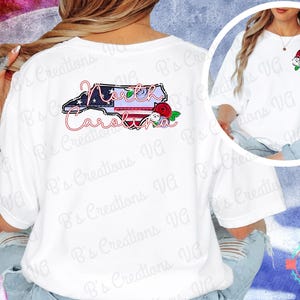Puede incluir: Camiseta blanca con un gráfico del contorno del estado de Carolina del Norte, que incorpora un diseño patriótico y elementos florales. El diseño incluye el nombre del estado. Es una prenda informal.