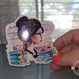 May include: A sticker featuring a woman with glasses and a bun, surrounded by books and flowers. The design includes a stack of blue books and a floral arrangement. The sticker has a glittery, holographic effect.