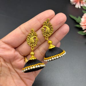 May include: A pair of black and gold jhumka earrings. The earrings feature a peacock design at the top and a bell-shaped bottom. The bottom of the earrings are adorned with white and yellow crystals. The earrings are held in a hand.