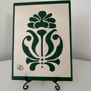 Puede incluir: Un azulejo de cerámica verde y blanco con un diseño floral estilizado. El azulejo está en un caballete negro.