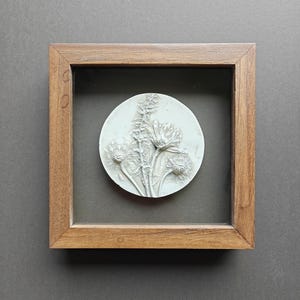 Op de afbeelding: Een vierkante houten lijst met een ronde witte reliëfplaat. De plaat is voorzien van een verhoogd, gedetailleerd botanisch ontwerp van drie bloemen met gestructureerde bloemblaadjes en stengels. Het kunstwerk is gecentreerd in de lijst.