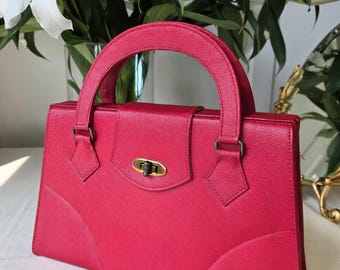 Può includere: Una borsa a mano in pelle rosa brillante con manico superiore e chiusura dorata. La borsa ha una forma strutturata e un davanti leggermente curvo.