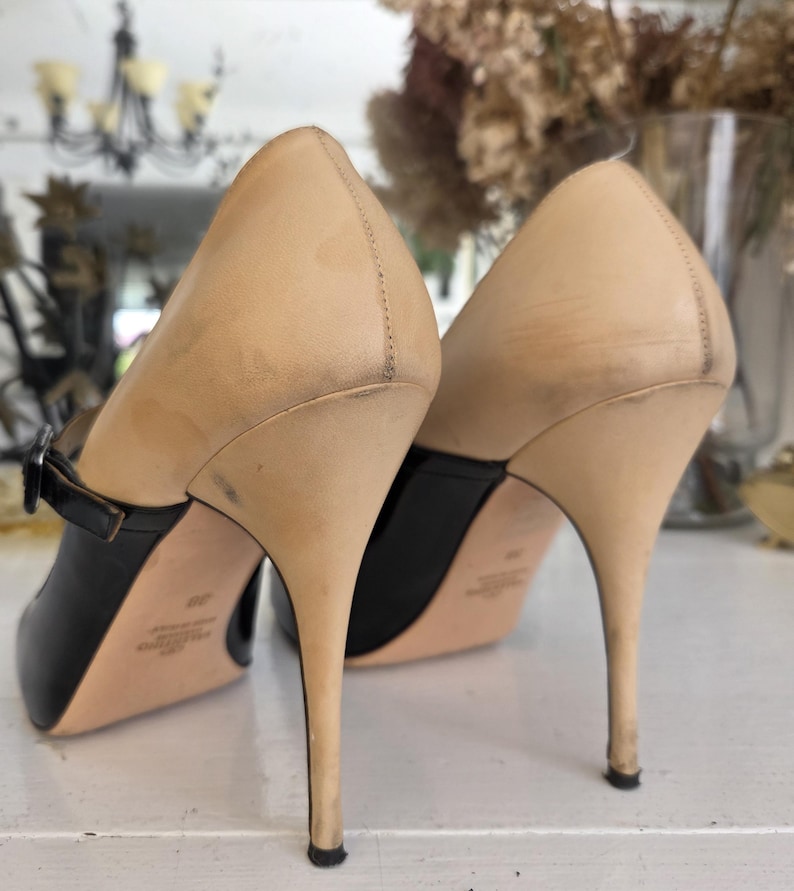 May include: A pair of high-heeled shoes with a black toe and strap, and a tan upper. The heels are slender and pointed. The shoes are displayed on a white surface.
