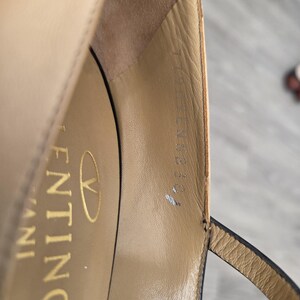May include: Close-up of a beige leather sandal with a thin ankle strap. The interior of the shoe features the brand name "Valentino" in gold lettering. The sandal has a high heel and a delicate design.