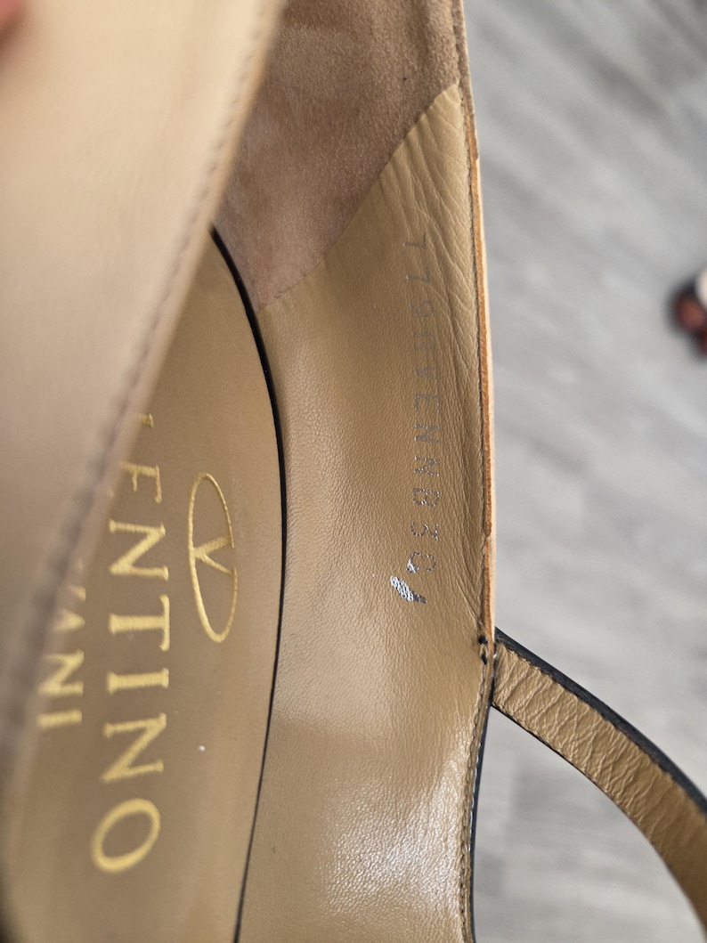 May include: Close-up of a beige leather sandal with a thin ankle strap. The interior of the shoe features the brand name "Valentino" in gold lettering. The sandal has a high heel and a delicate design.